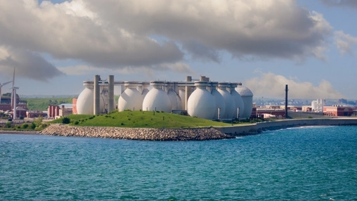 White spherical storage tanks on a green hillside by the sea with a wind turbine and industrial buildings