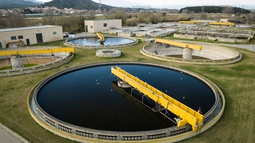 Dark circular tank with a yellow skimmer bridge sits among smaller treatment tanks backed by distant mountains