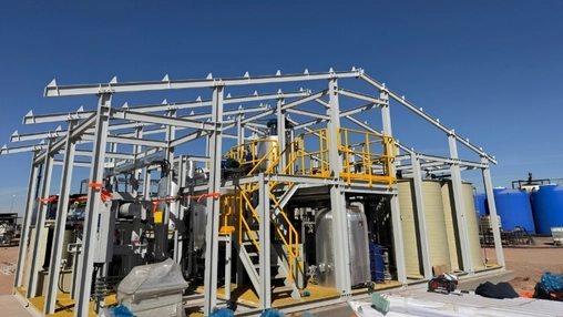 Open steel frame housing processing equipment with yellow safety railings and blue tanks in a desert setting