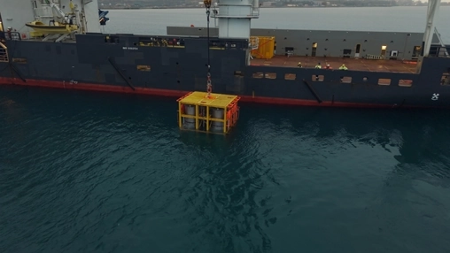 A yellow subsea unit in a metal frame is lowered by crane from a large support vessel into coastal water